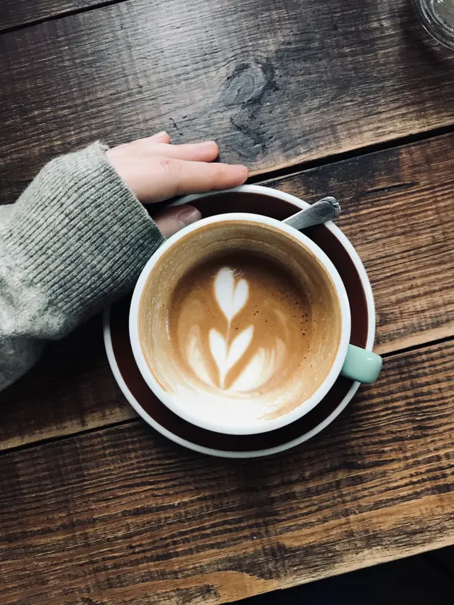 overhead-vertical-shot-person-s-hand-near-latte-art-coffee-wooden-surface.jpg